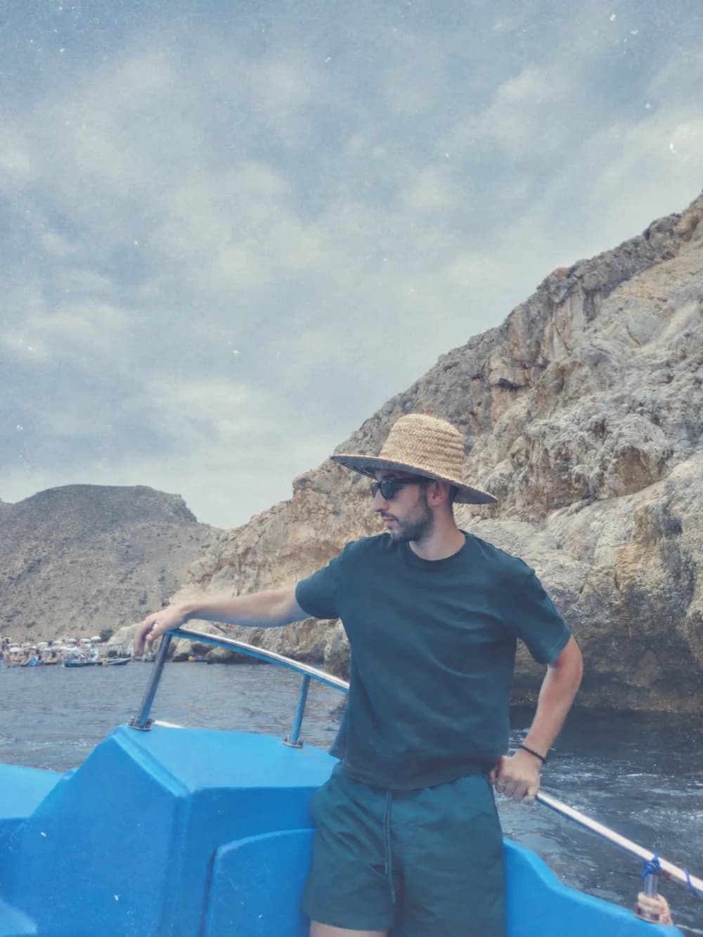Akram Tissi at the bow of a blue boat, in a straw hat, looking out at a rock face.